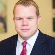 A white man with short brown hair whearing a dark suit, white shirt and red tie.