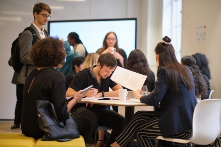 group of students looking at papers around a table, with more students in the background