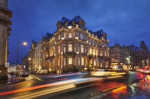 Illuminated historic hotel building on a city street at dusk, with blurred vehicle light trails reflecting on the wet road.