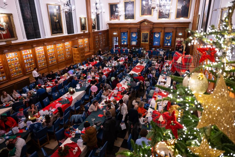 Overhead view of a festive Christmas event in a historic hall, with families seated at decorated tables and a large Christmas tree adorned with lights and ornaments in the foreground.