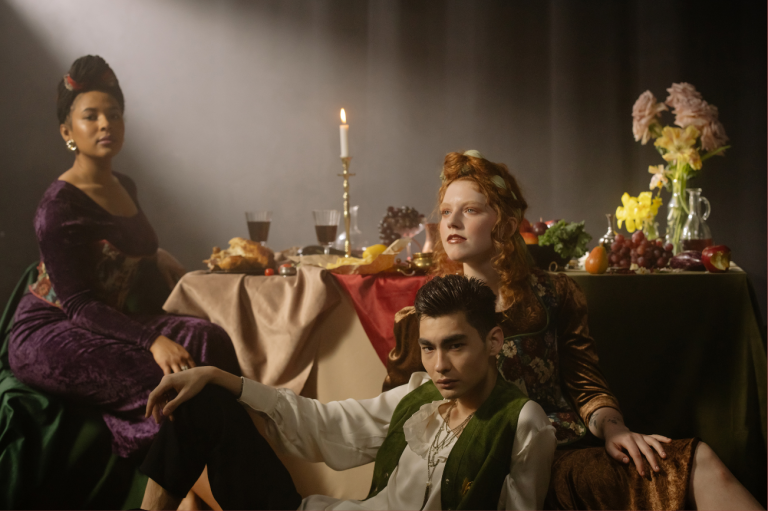 Two women in a Renaissance-style setup with fruits, flowers, and a candlelit table.
