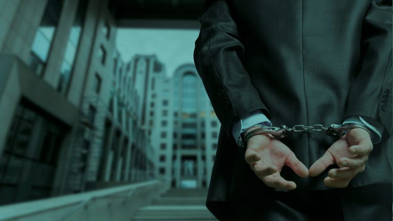 Person in a dark business suit stands with hands cuffed behind their back in an urban setting. The focus is on the metal handcuffs and restrained hands in the foreground, while modern office buildings and a walkway appear blurred in the background. The image has cool blue-green tones and a dramatic, low-angle perspective.