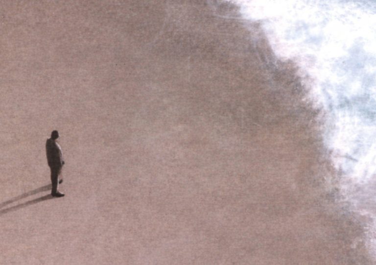 Aerial view of a large sandy beach with a single person standing near the water’s edge. White sea foam from incoming waves stretches along the right side of the image, contrasting with the smooth, pale sand. The person appears small within the wide open landscape, casting a long shadow across the beach. The composition emphasizes scale, space, and the relationship between the individual and the natural environment. For EDI contexts, the image can be interpreted as representing inclusion and belonging through the presence of an individual within a shared public space, without specifying personal characteristics.