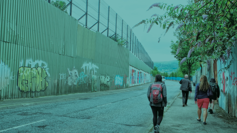 A group of pedestrians walk along a quiet street bordered by tall corrugated metal walls covered in graffiti and posters. A high security fence runs along the top of the wall on the left, while flowering trees line the right side of the road. Hills are visible in the distance under an overcast sky.