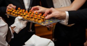A server wearing white gloves offers a tray of bite-sized canapés to guests at a reception. Several guests are reaching for the food.