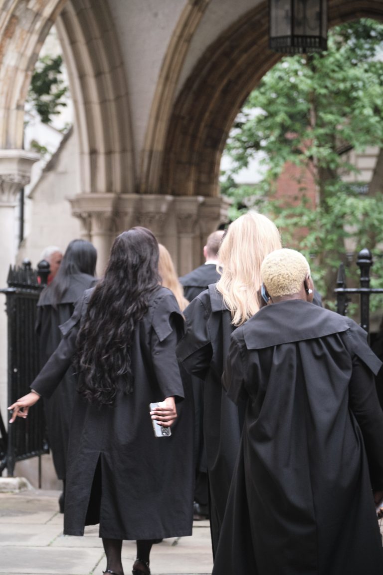 Students in gowns photographed from behind, proceeding in line through Church Court