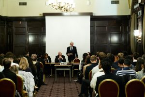 Speaker addressing an audience during a panel event in a conference room with two seated panellists and a large projection screen.