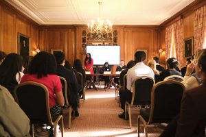 Presenter addressing an audience during a panel discussion in a historic meeting room with a large screen and seated panellists.