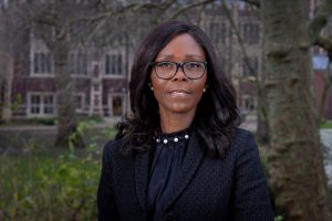 Professional head-and-shoulders outdoor portrait of an individual wearing dark-framed glasses, a black textured blazer, and a high-neck black blouse with pearl detailing. The individual stands facing the camera in a landscaped setting with trees, greenery, and a blurred historic-style building in the background. Natural daylight creates soft, even lighting across the scene. The image presents a professional and inclusive representation, including a visible glasses wearer, within a formal leadership-style portrait.