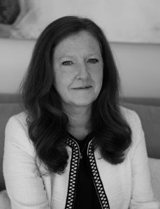 Black-and-white head-and-shoulders portrait of a person with long hair, seated indoors and facing the camera. The person is wearing a light-coloured textured jacket with dark patterned trim over a dark top. The background is softly blurred, showing part of a sofa and wall artwork. Soft, even lighting highlights facial features and hair, creating a professional and formal portrait style.