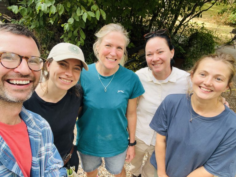 Group selfie taken outdoors showing five people standing close together in a garden setting. One person in the foreground holds the camera while the others gather around, all looking toward the lens. The group is dressed casually, including T-shirts, a light shirt, and a baseball cap. Trees and leafy shrubs fill the background, with gravel underfoot and garden furniture partially visible in the distance. Natural daylight illuminates the scene.