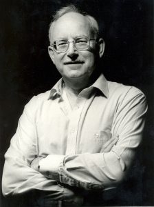 Black-and-white studio portrait of a person wearing glasses and a light-coloured button-down shirt. The person is standing with arms folded across the chest, facing the camera. The image is tightly framed from about the waist up, with dramatic lighting that highlights the face and shirt against a dark, plain background.