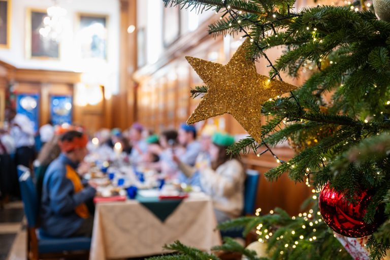 A decorated Christmas tree fills the foreground, featuring a glittering gold star ornament, a red bauble, and warm white fairy lights. In the softly blurred background, people are seated at long tables in a wood-panelled hall, gathered for a festive meal or holiday event.