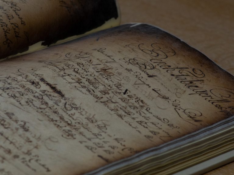 Close-up of an open antique manuscript or ledger with aged, yellowed pages covered in ornate handwritten calligraphy. The pages have darkened, worn edges and are filled with flowing cursive writing in faded black ink, photographed at an angle under soft, warm lighting.