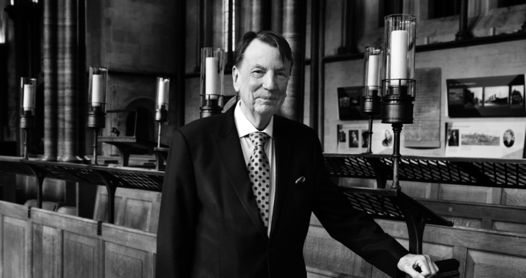 Black-and-white photograph of a person standing inside a large historic interior with stone columns, wooden panelling and candle-style light fixtures. The person is wearing a dark suit, a light-coloured shirt and a polka-dot tie, with one hand resting on a wooden rail. Framed displays and photographs are visible on the walls in the background. The image is professionally composed, with the subject positioned centrally and lit by soft indoor lighting.