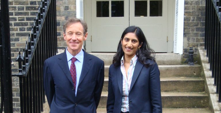 Two people in business attire stand side by side on stone steps outside a brick building with a white entrance door and black railings. One person is wearing a dark suit with a light blue shirt and patterned tie, while the other is wearing a dark blazer over a striped shirt. The photograph is taken outdoors in daylight, with the building entrance forming a symmetrical backdrop.