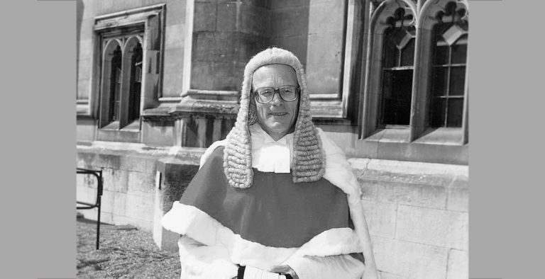 Black-and-white photograph of a person standing outdoors in front of a historic stone building with Gothic-style windows. The person is wearing a traditional judicial wig, glasses and formal ceremonial robes trimmed with fur. The image is centred on the figure, with architectural details of the building visible in the background.
