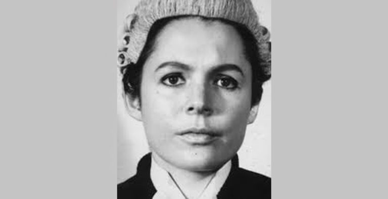 Black-and-white close-up portrait of a person wearing a traditional judicial wig and formal court dress. The person is facing directly towards the camera, with the image tightly cropped around the head and shoulders. The background is plain and softly lit, drawing attention to the facial features and the textured curls of the wig. The photograph has a formal, professional style commonly associated with legal or judicial portraits.
