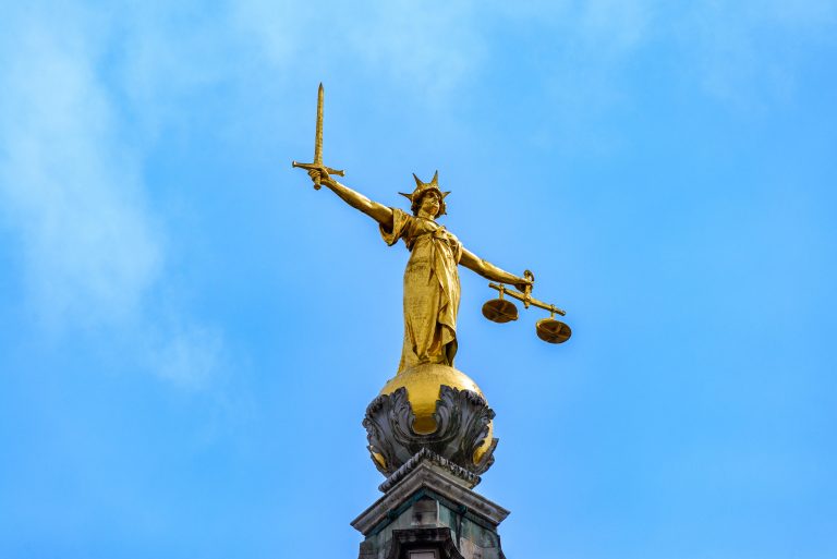 Looking up at a gold statue of lady justice with a blue sky with light cloud