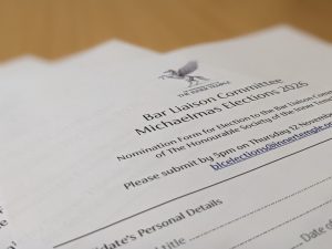 Close-up, angled photograph of a printed nomination form from The Honourable Society of the Inner Temple titled “Bar Liaison Committee Michaelmas Elections 2026.” The document is placed on a light wooden surface, with the centre text in focus and the edges softly blurred. Visible text includes instructions to submit nominations by 5pm on Thursday 12 November and an elections email address. A “Candidate’s Personal Details” section with blank lines appears lower on the page. The lighting is soft and warm, creating a professional, document-focused image.