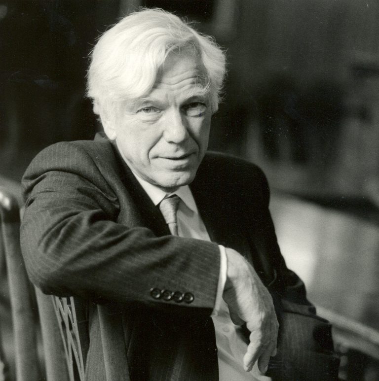 Black-and-white portrait of a person with light-coloured hair, wearing a dark suit, light shirt and tie, seated sideways on a decorative wooden chair. One arm rests over the chair back while the other hand rests near the lap. The photograph is closely framed, with a softly blurred indoor background that draws attention to the subject.