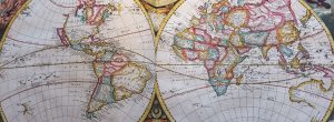 Vintage double-hemisphere world map showing the Americas on the left and Europe, Africa, Asia and Australia on the right. Countries and regions are outlined in pastel shades of pink, blue, yellow and green, with fine black place names, latitude and longitude gridlines, and decorative celestial illustrations between the two circular map panels. The map has an aged paper texture and historical cartographic style.