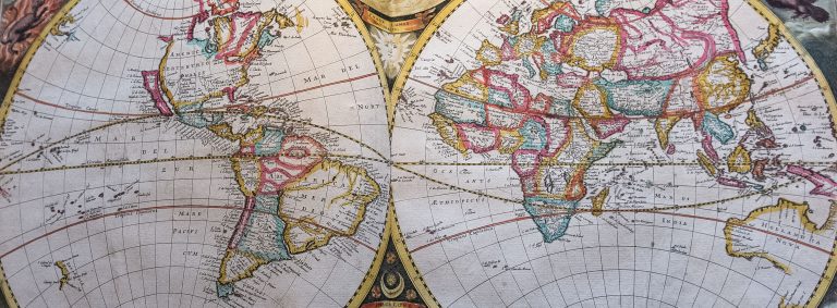 Vintage double-hemisphere world map showing the Americas on the left and Europe, Africa, Asia and Australia on the right. Countries and regions are outlined in pastel shades of pink, blue, yellow and green, with fine black place names, latitude and longitude gridlines, and decorative celestial illustrations between the two circular map panels. The map has an aged paper texture and historical cartographic style.