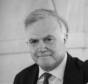 Black-and-white portrait of a person in a dark business suit and patterned tie, leaning an arm against a stone pillar. The person is positioned in the foreground and looking toward the camera. The background features an indoor architectural setting with curved stone archways and masonry details, softly out of focus.