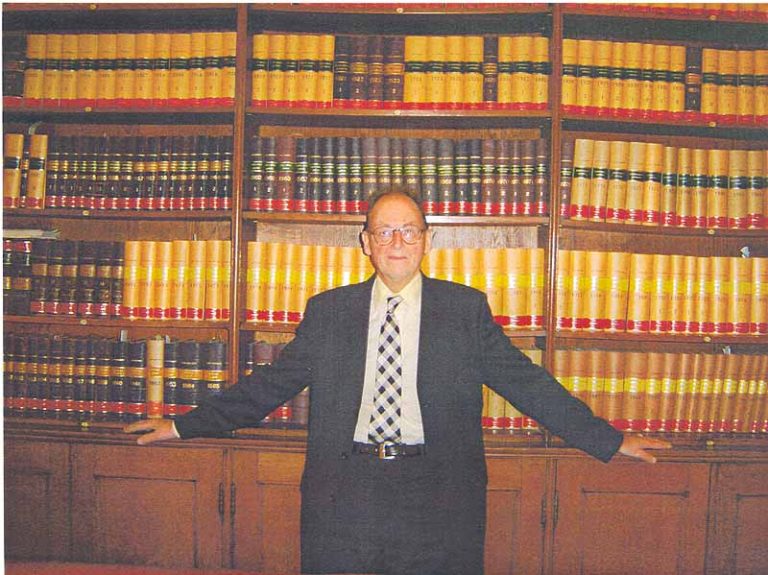 A person wearing a dark suit, light shirt and patterned tie stands in front of large wooden bookshelves filled with rows of bound legal-looking volumes. The person is positioned centrally with arms resting on the shelving units on either side. The room has warm lighting and a traditional library or archive setting.